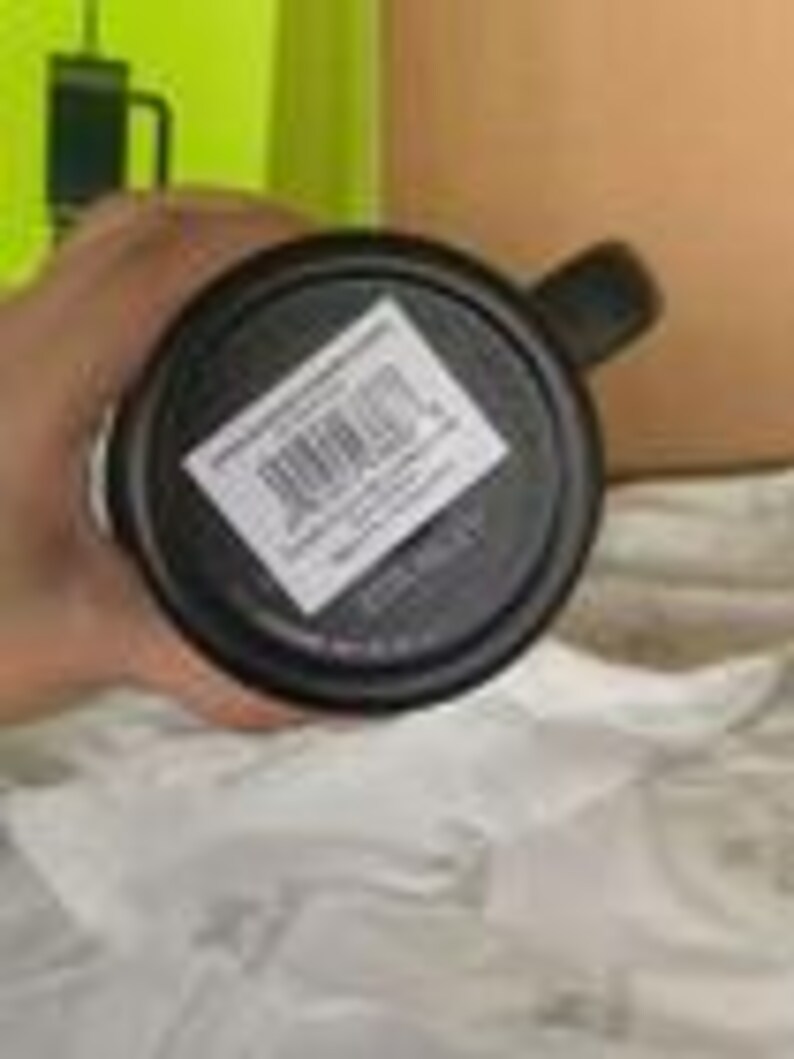Peut inclure: Un mug noir avec une anse, vu par le dessous. Une &eacute;tiquette rectangulaire blanche est appos&eacute;e sur la base, pr&eacute;sentant un code-barres, le texte 'MADE IN CHINA' et d'autres petits d&eacute;tails imprim&eacute;s. Le mug a une base circulaire et une finition sombre.