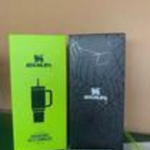 Peut inclure: Une bo&icirc;te de produit rectangulaire vert citron vif pr&eacute;sentant un graphique noir d'un gobelet Stanley avec une paille et une anse, &agrave; c&ocirc;t&eacute; d'une bo&icirc;te rectangulaire noire avec un motif sombre subtil et le logo Stanley blanc centr&eacute; sur la face avant.