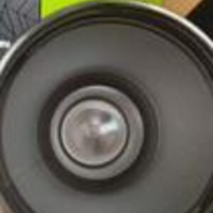 Peut inclure: Un haut-parleur circulaire gris fonc&eacute; dot&eacute; d'un cache-noyau central en m&eacute;tal argent&eacute;. La membrane du haut-parleur est lisse et mate, entour&eacute;e d'un anneau ext&eacute;rieur &eacute;pais en caoutchouc noir. Une bo&icirc;te vert citron avec le logo Stanley est visible.