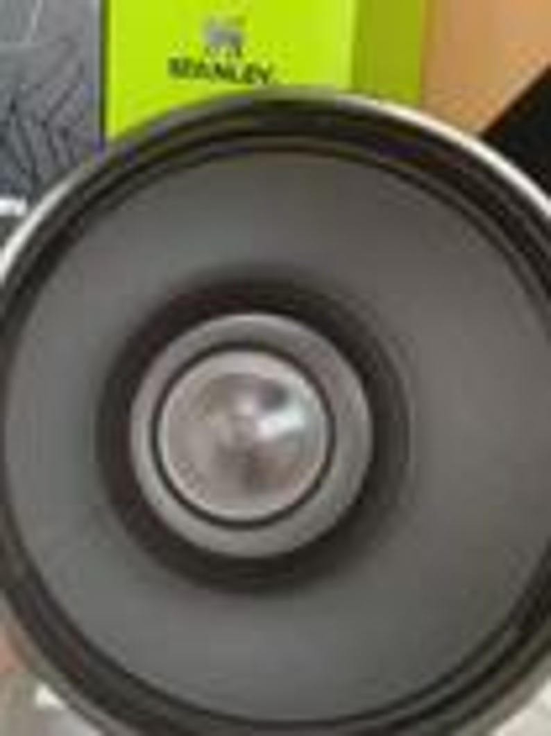 Peut inclure: Un haut-parleur circulaire gris fonc&eacute; dot&eacute; d'un cache-noyau central en m&eacute;tal argent&eacute;. La membrane du haut-parleur est lisse et mate, entour&eacute;e d'un anneau ext&eacute;rieur &eacute;pais en caoutchouc noir. Une bo&icirc;te vert citron avec le logo Stanley est visible.
