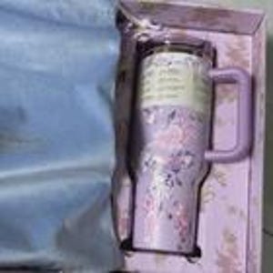 Pu&ograve; includere: Un bicchiere in acciaio inossidabile color lavanda con manico e coperchio trasparente, caratterizzato da un motivo floreale nei toni del rosa e del viola. Il bicchiere &egrave; contenuto in una scatola color lavanda con rivestimento in tessuto azzurro.