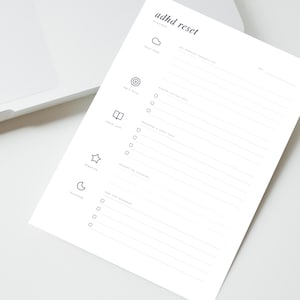 Könnte beinhalten: Ein weißer Planer mit dem Titel "adhd reset" mit minimalistischem Design. Der Planer enthält Abschnitte für Brain Dump, Top 3 Fokus, Schulaufgaben, Träumen, Launchpad und andere Aufgaben. Der Planer liegt auf einer weißen Oberfläche.