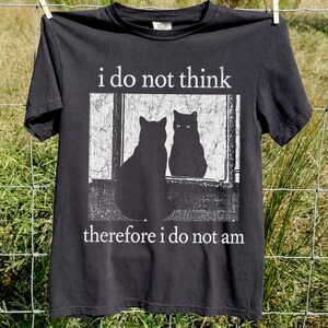 May include: A charcoal gray short-sleeve t-shirt featuring a white graphic of a cat sitting in front of a mirror. The text above the graphic reads i do not think and the text below the graphic reads therefore i do not am in white font.