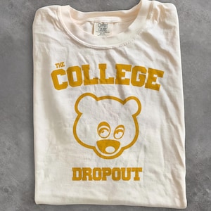 May include: A cream-colored short-sleeve t-shirt featuring a gold graphic of a bear head centered on the chest. The text "THE COLLEGE DROPOUT" is printed in gold block lettering above and below the bear. A Comfort Colors brand tag is visible.