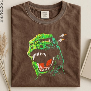May include: An espresso-colored t-shirt featuring a graphic print of a green monster with an open mouth showing white teeth and a red tongue. Two white fighter jets with orange exhaust trails are positioned above the monster's head.