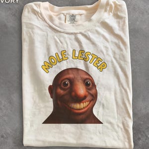 May include: An ivory-coloured t-shirt featuring a graphic of a brown, bald, smiling character with large eyes and prominent teeth. Above the character, the text MOLE LESTER is printed in yellow letters. The shirt has a crew neckline and a Comfort Colors label.