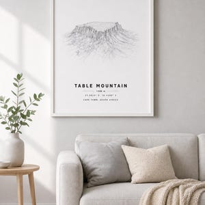 Könnte beinhalten: Ein gerahmter Kunstdruck mit einer schwarz-weißen Strichzeichnung des Tafelbergs. Der Text lautet TABLE MOUNTAIN, 1086 m, 33.9628 S, 18.4098 E und CAPE TOWN, SOUTH AFRICA. Der Druck befindet sich in einem schlichten weißen Rahmen an der Wand.