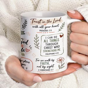 May include: A white ceramic mug featuring black printed text and botanical illustrations. The text includes Bible verses such as Trust in the Lord with all your heart, I can do all things through Christ who strengthens me, and For we walk by faith.