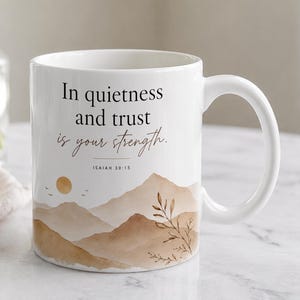 May include: A white ceramic mug featuring the black text In quietness and trust is your strength. Isaiah 30:15. The design includes a watercolor-style illustration of tan and brown mountains, a golden sun, and a leafy branch.