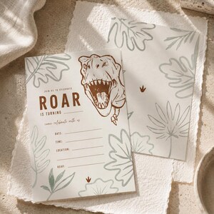 Könnte beinhalten: Ein Satz Einladungskarten zum Thema Dinosaurier. Die Hauptkarte zeigt eine braune Tyrannosaurus Rex-Illustration und den Text "ROAR IS TURNING". Das Design umfasst grüne Blatt- und Farnakzente. Der passende Umschlag hat ein ähnliches Design.