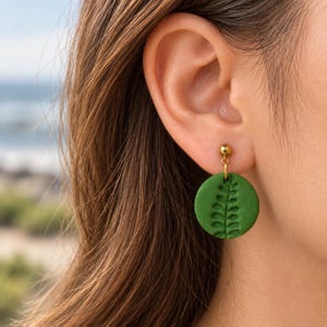 Peut inclure: Une boucle d'oreille circulaire verte avec un motif de feuille de fougère en relief. Le disque est suspendu à un petit clou d'oreille doré en forme de bille. Le design est plat et rond, présentant une texture botanique détaillée pressée.