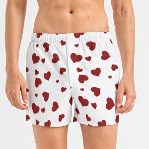 May include: White boxer shorts patterned with red hearts. The shorts feature an elasticated waistband and a button closure. The hearts are of varying sizes and are spread across the fabric.
