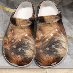 May include: A pair of slip-on clogs featuring a realistic brown and black fur print pattern. The footwear has a white interior lining, a pivoting heel strap, and multiple circular ventilation holes across the top and sides of the rounded toe area.