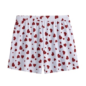 May include: White athletic shorts featuring an all-over print of small, scattered dark red hearts. The shorts have a wide, elasticated waistband with a gathered texture and a straight-cut hemline, providing a simple and functional design.