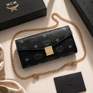 May include: Black leather wallet with a gold chain strap. The wallet features a repeating pattern and a gold-coloured clasp. The MCM logo is visible on the wallet, box, and card.