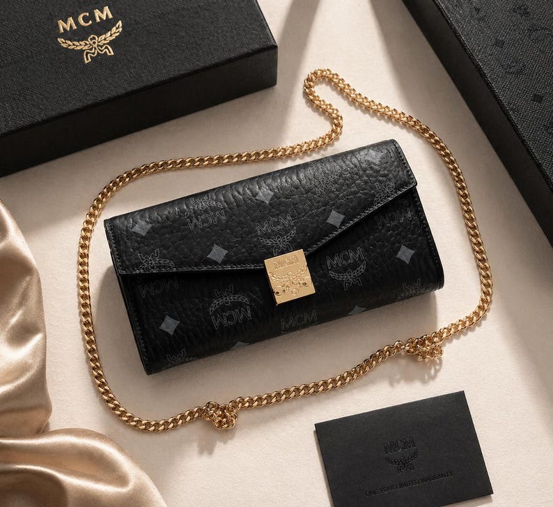 May include: Black leather wallet with a gold chain strap. The wallet features a repeating pattern and a gold-coloured clasp. The MCM logo is visible on the wallet, box, and card.