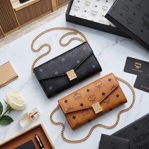 May include: Two MCM crossbody wallets in black and tan leather with gold chain straps. The wallets feature the MCM logo pattern and gold hardware. Accessories include a gold card case, perfume, and a white rose.