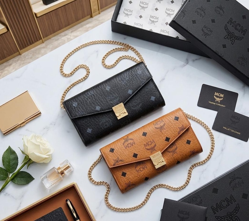 May include: Two MCM crossbody wallets in black and tan leather with gold chain straps. The wallets feature the MCM logo pattern and gold hardware. Accessories include a gold card case, perfume, and a white rose.