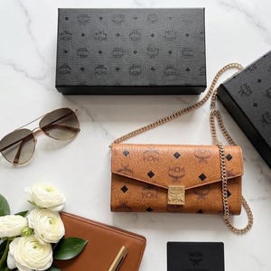 May include: A tan leather wallet-style crossbody bag with a gold chain strap, featuring a repeating logo pattern and a gold clasp. Also, a pair of aviator sunglasses, a brown leather wallet, and a black box.