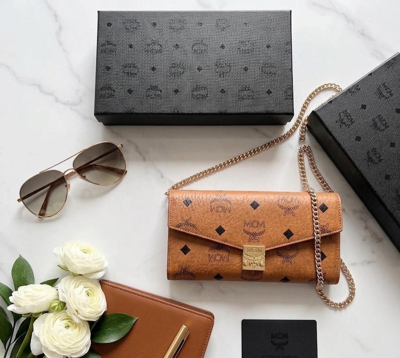 May include: A tan leather wallet-style crossbody bag with a gold chain strap, featuring a repeating logo pattern and a gold clasp. Also, a pair of aviator sunglasses, a brown leather wallet, and a black box.