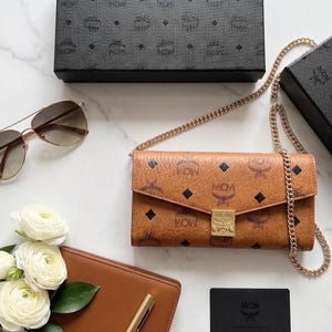 May include: A brown MCM crossbody wallet with a gold chain strap, featuring the MCM logo pattern. The image also includes a pair of aviator sunglasses, a brown leather wallet, white flowers, and black boxes with the MCM logo.