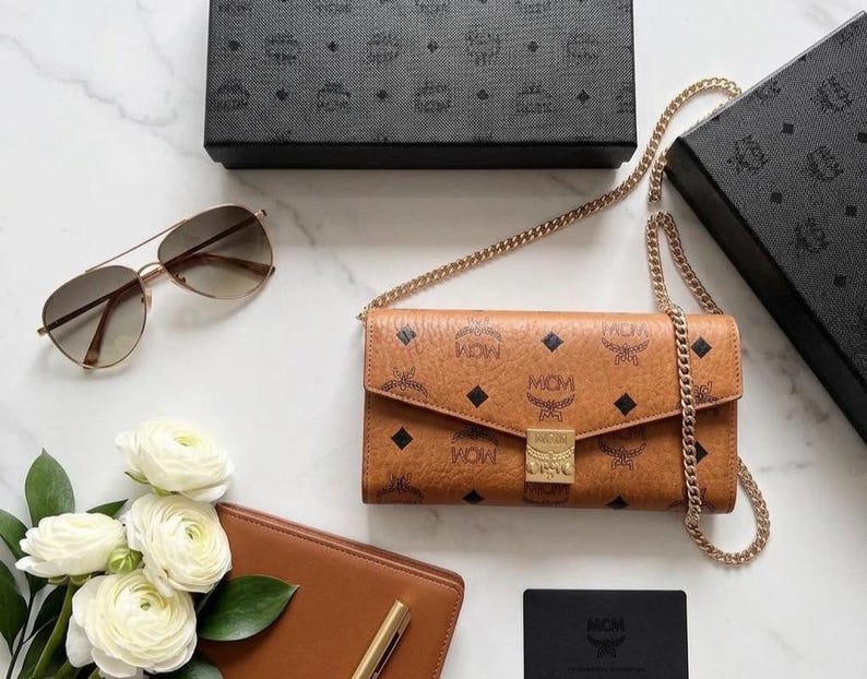 May include: A brown MCM crossbody wallet with a gold chain strap, featuring the MCM logo pattern. The image also includes a pair of aviator sunglasses, a brown leather wallet, white flowers, and black boxes with the MCM logo.