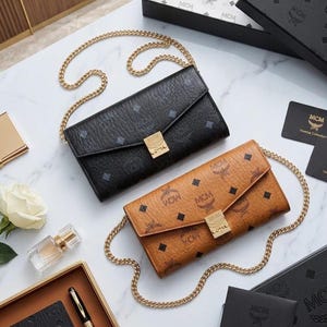May include: Two MCM wallets with gold chain straps are displayed on a marble surface. One is black, the other is tan, both with a repeating logo pattern. Additional accessories include a gold card holder, perfume, and a white rose.