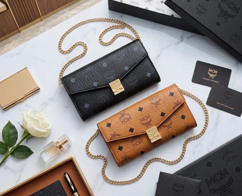 May include: Two MCM wallets with gold chain straps are displayed on a marble surface. One is black, the other is tan, both with a repeating logo pattern. Additional accessories include a gold card holder, perfume, and a white rose.