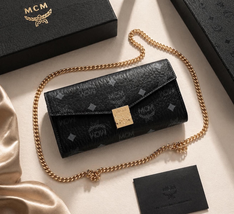May include: Black leather wallet with a gold chain strap. The wallet features a repeating pattern and a gold-coloured clasp. The MCM logo is visible on the wallet, box, and card.
