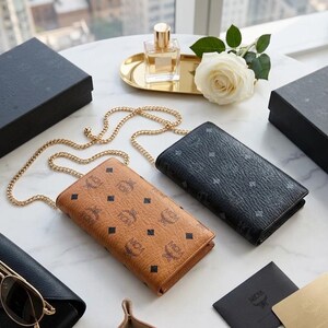 May include: Two leather wallets with gold chain straps, one tan and one black, are displayed on a marble surface. The tan wallet has a repeating logo pattern with black diamond accents. A pair of sunglasses and a perfume bottle are also visible.