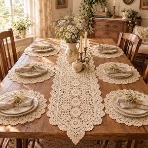 Könnte beinhalten: Ein cremefarbener Häkel-Tischläufer mit wiederkehrendem Blumenmuster und gewelltem Rand, begleitet von passenden runden Häkel-Platzdeckchen. Das Set ist auf einem hölzernen Esstisch mit weißen Keramiktellern und Leinenservietten arrangiert.