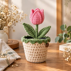 Op de afbeelding: Een gehaakte roze tulp met groene bladeren die uit een crèmekleurige gehaakte pot komt. De pot heeft een geschulpte groene bovenrand en een kleine bruine ronde knoop aan de zijkant. Het geheel is volledig gemaakt van garen.