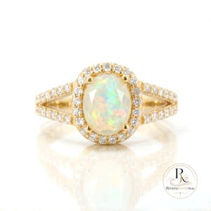 May include: A gold ring featuring an oval opal gemstone surrounded by a halo of small diamonds. The ring's band splits into two diamond-studded sections on each side of the opal, creating a split-shank design. The ring is set against a white background.