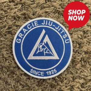 May include: A circular blue and white patch with the words "GRACIE JIU-JITSU" around the edge and "SINCE 1925" at the bottom. The center features a white triangle with a stylized illustration of two figures in a grappling position. A red "SHOP NOW" speech bubble is in the upper right.