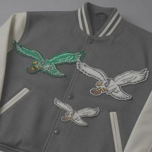 May include: Gray varsity jacket with white sleeves and collar. The jacket features embroidered patches of eagles holding footballs in green and white. The jacket has a button-up front.