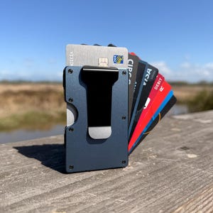 Peut inclure: Un porte-cartes en métal bleu foncé doté d'une pince à billets noire sur le devant. Le portefeuille est déployé pour afficher plusieurs cartes de crédit et de débit, dont une carte argentée avec le texte RBC et une carte rouge avec le texte DEBIT.