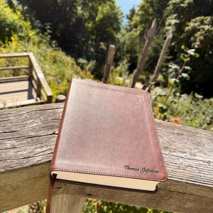 Op de afbeelding: Een bruin, in leer gebonden boek met witte stiksels langs de randen. De kaft is voorzien van een klein gouden kruissymbool in de rechterbovenhoek en de naam Thomas Jefferson in zwarte schrijfletters in de rechterbenedenhoek met een leeslint.