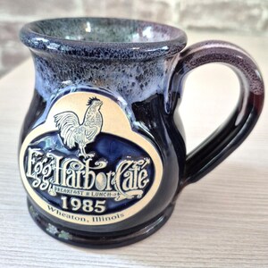 May include: A dark blue ceramic mug with a glossy finish and a lighter blue rim. The front features a raised oval emblem with a rooster illustration, the text Egg Harbor Cafe, Breakfast & Lunch, 1985, and the location Wheaton, Illinois.