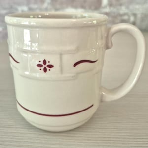 May include: A cream-colored ceramic mug featuring a raised basket-weave texture around the upper portion. The mug is decorated with a small, dark red four-petaled floral motif and thin, horizontal dark red lines encircling the body.
