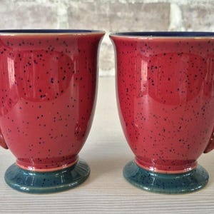 May include: Two red ceramic mugs with speckled patterns and dark blue rims and bases. The mugs have large handles and a glossy finish, perfect for coffee or tea.