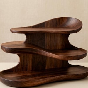 Può includere: Un espositore a tre livelli in legno marrone scuro con ripiani lisci e ondulati a forma di onda. La struttura è realizzata in legno con venature visibili e presenta tre livelli distinti per il posizionamento, ciascuno con una forma curva.