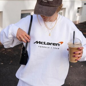 Può includere: Una felpa bianca a maniche lunghe con girocollo che presenta il logo McLaren nero con un arco arancione e la scritta Formula 1 Team stampata in nero sul petto. La felpa è abbinata a un cappellino da baseball marrone scuro e a una borsa nera.