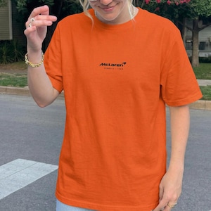 Può includere: Una t-shirt a maniche corte di colore arancione brillante con la scritta nera McLaren Formula 1 Team stampata al centro del petto. Il capo presenta un girocollo e una vestibilità standard, offrendo un look semplice e pulito per l'uso quotidiano.