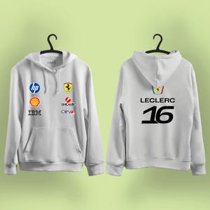 Może przedstawiać: Biała bluza z kapturem z wieloma logo sponsorów z przodu, w tym HP, Shell i IBM. Z tyłu napis "LECLERC" i numer "16" w kolorze czarnym. Widoczne jest również logo Ferrari.