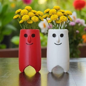 May include: Two cylindrical vases, one red and one white, each featuring a carved face with circular eyes, a triangular nose, and a curved mouth. Both vases are filled with bright yellow button flowers, standing upright on a dark surface.
