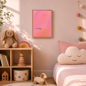 Könnte beinhalten: Ein rechteckiger Wandkunstdruck in einem hellen Holzrahmen mit einem rosa-orangen Farbverlauf und dem schwarzen Text "DU WIRST GELIEBT, LITTLE J." in der unteren Mitte des Drucks, präsentiert an einer einfarbigen hellrosa Wand.