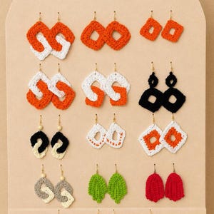 May include: A display of handmade crochet earrings in various shapes and colors. The earrings include orange and white, black, green, and red designs. Some earrings have wooden bead accents. The earrings are displayed on a neutral-toned background.