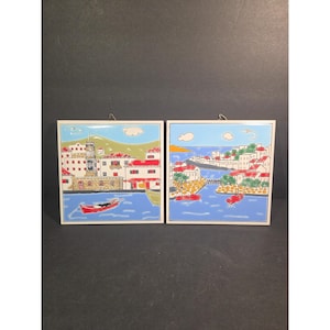 Può includere: Due piastrelle quadrate in ceramica con scene costiere dipinte. Ogni piastrella presenta un cielo blu, acqua, barche rosse ed edifici bianchi con tetti rossi. Una piastrella ha un campanile. Le piastrelle sono incorniciate in bianco.