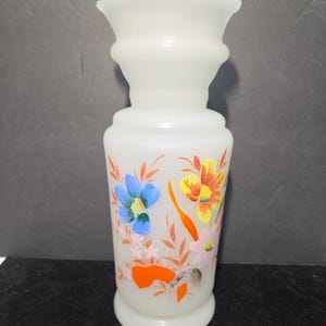 May include: A white milk glass vase with a scalloped rim and a cylindrical body. The exterior features a hand-painted floral design with blue, yellow, orange, and pink flowers accented by thin orange leaves and stems against the opaque glass.
