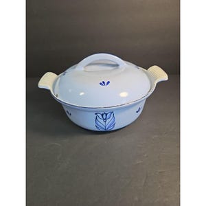May include: A light blue ceramic casserole dish with a lid and handles. The dish features a floral design in shades of blue. The handles and the handle on the lid are white. The dish is round and appears to be vintage.